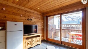 Een houten interieur met een televisie op een houten kast, een koelkast en een schuifdeur naar een buitenruimte met een tafel en stoelen.
