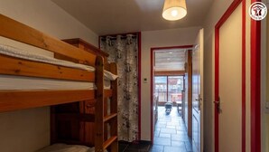Een smalle gang met een houten stapelbed aan de linkerkant, een kamerbreed gordijn en een deur naar een balkon.