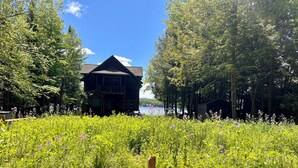 A wooden cabin with a balcony, surrounded by lush greenery and a lake. - 4th Lake Getaway, Screened Porch, Large Deck & Private Dock (Old Forge)