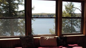 A cabin with a large window overlooking a lake, a wooden bench with cushions, and a view of trees and hills. - 4th Lake Getaway, Screened Porch, Large Deck & Private Dock (Old Forge)