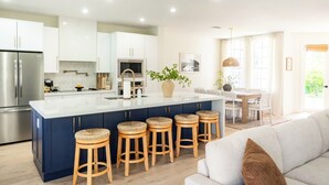 A modern kitchen with white cabinets, a stainless steel refrigerator, a large island with stools, and a dining area with a table and chairs. - NEW Luxe Wellness Retreat - Private Gym, Pilates, Sauna, Pool (Delray Beach)