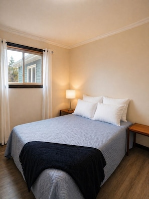 A bedroom with a bed, a nightstand, a lamp, a window with curtains, and a view of a house outside.