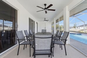 Un espace de restauration extérieur abrité, avec vue sur la piscine, comprenant une table et des chaises, des ventilateurs de plafond et de grandes fenêtres.
