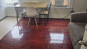 Una habitación con piso de madera, una mesa de comedor con sillas, un sofá gris y una ventana con persianas.