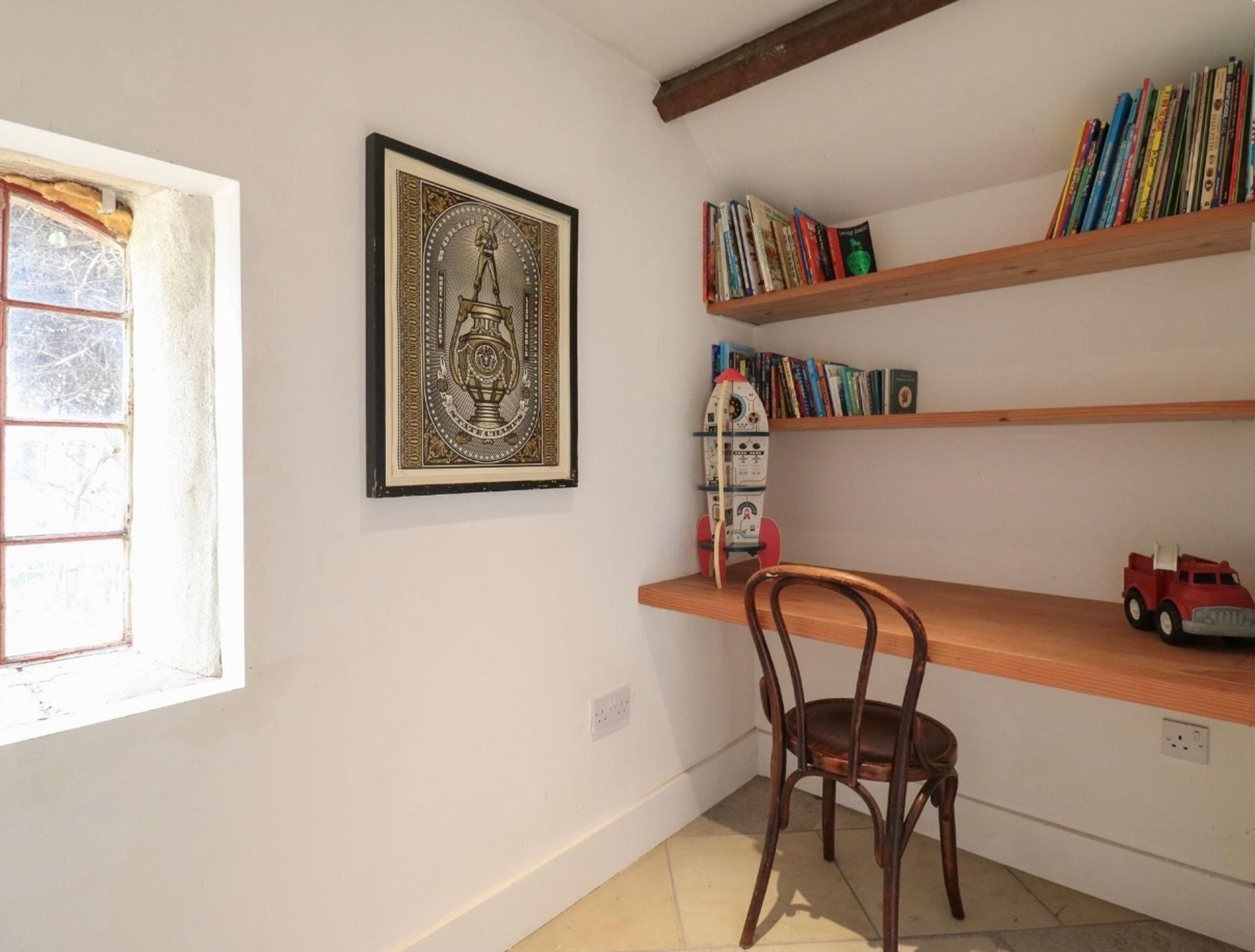A room with a wooden chair, a bookshelf with books, a framed picture, and a window with a view of trees.