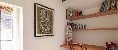 A room with a wooden chair, a bookshelf with books, a framed picture, and a window with a view of trees.