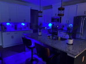 Una cocina moderna con gabinetes blancos, encimera de granito e iluminación azul debajo de los armarios. Hay dos taburetes de bar y dos plantas en macetas en la isla.