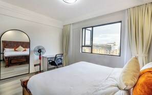 A hotel room with a bed, a desk with a chair, a standing fan, a window with a view of buildings, and a mirror on the wall. - Riara one residency (Nairobi)