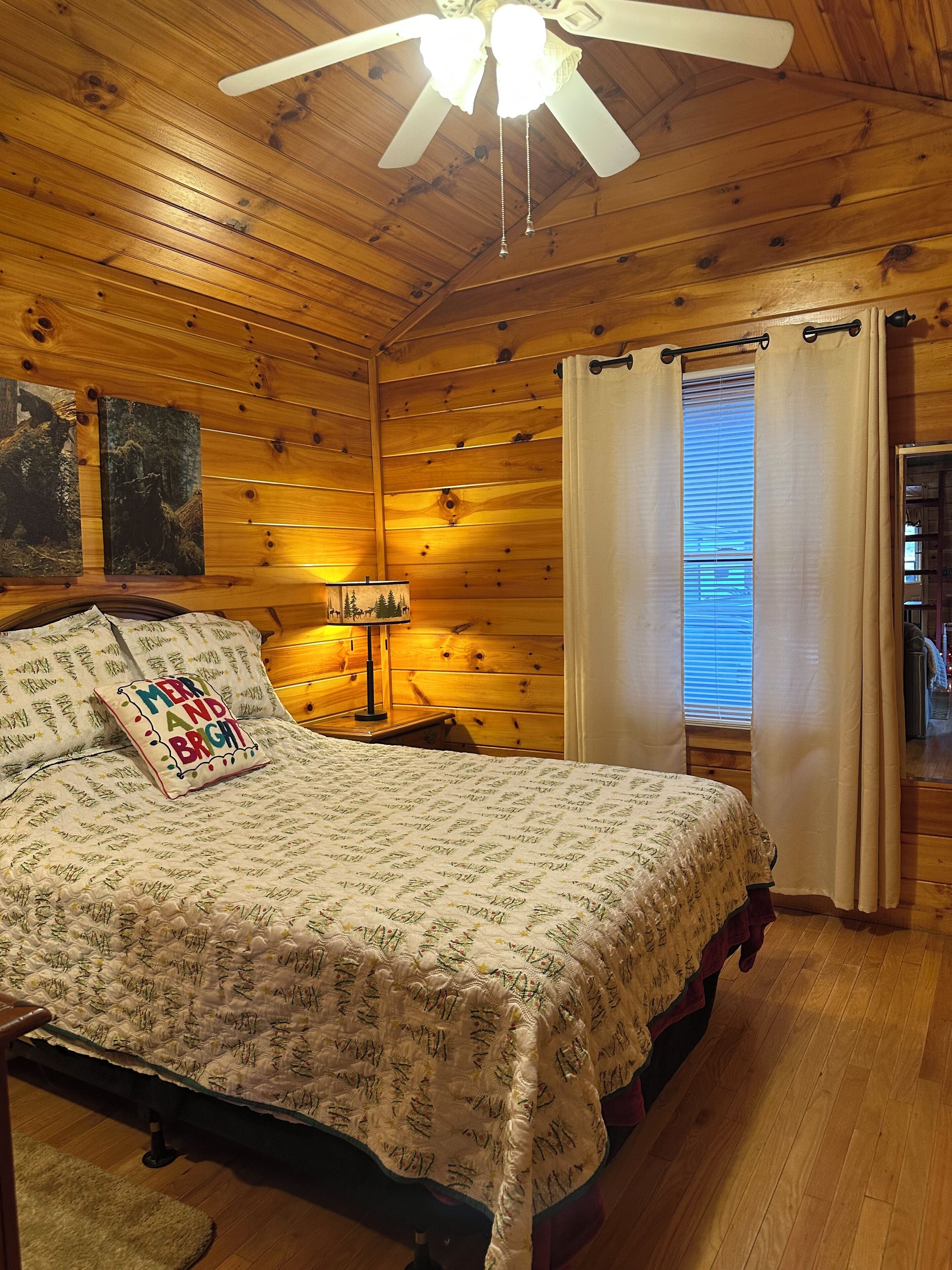 A cozy wooden cabin bedroom with a bed, a ceiling fan, a nightstand with a lamp, and a window with curtains.