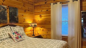 Accogliente camera da letto in una baita di legno, con un letto, una ventola a soffitto, un comodino con una lampada e una finestra con tende.
