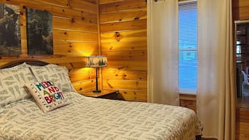 A cozy wooden cabin bedroom with a bed, a ceiling fan, a nightstand with a lamp, and a window with curtains.