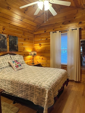 A cozy wooden cabin bedroom with a bed, a ceiling fan, a nightstand with a lamp, and a window with curtains. - Riverfront Cabin Fishing extravaganza! Family Friendly! (Bryson City)