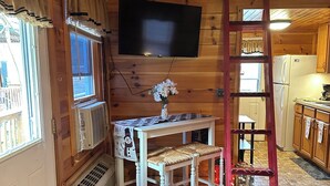Un accogliente interno di una baita con arredamento in legno, una televisione montata a parete, un tavolo da pranzo con sedie, una scala e un frigorifero nella zona cucina.
