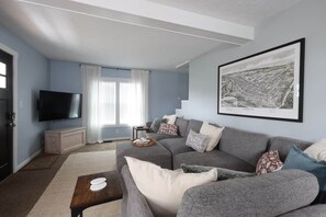 Una sala de estar con un sofá gris, un televisor de pantalla plana, una obra de arte con una vista urbana enmarcada y una ventana con cortinas.