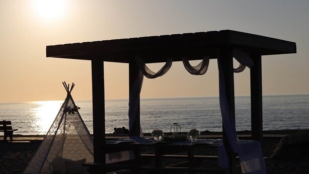 Cena de praia com um gazebo de madeira, um tipi e uma mesa posta para refeição.