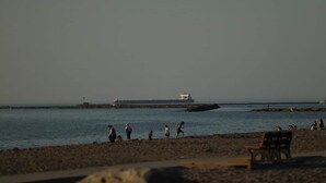 Uma praia com pessoas, um banco e um navio grande ao longe.