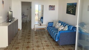 A living room with a sofa, a coffee table, and a TV on a stand. There is a dining area with chairs and a table, and a kitchen area with a fridge. - Fronte Mare 2 (Torre Lapillo)