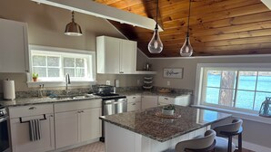 A kitchen with white cabinets, granite countertops, a central island with bar stools, a window with a view of the water, and a ceiling with exposed wooden beams. - Waterfront Family Getaway with Sunroom & Stunning Views (Washington)