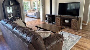 A living room with a leather sofa, a TV on a wooden stand, a fireplace, and a view through a glass door. - Waterfront Family Getaway with Sunroom & Stunning Views (Washington)