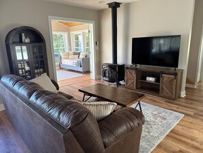 A living room with a leather sofa, a TV on a wooden stand, a fireplace, and a view through a glass door. - Waterfront Family Getaway with Sunroom & Stunning Views (Washington)