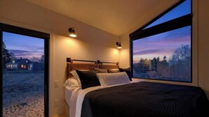 A bedroom with a large bed, two windows, and a view of a landscape. - Osprey Cabin - Ocean Front Acadia A Frame Cabin (Tremont)