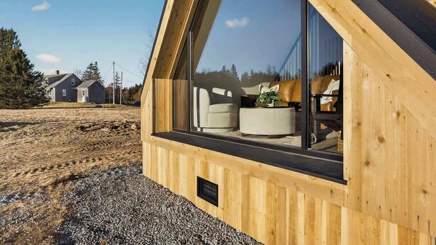 A modern wooden cabin with a large window, a visible interior with a sofa and a plant, and a gravel ground. - Osprey Cabin - Ocean Front Acadia A Frame Cabin (Tremont)