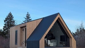 A wooden cabin with a triangular roof, a visible interior with a sofa and a dining area, and a small set of stairs leading to the entrance. - Osprey Cabin - Ocean Front Acadia A Frame Cabin (Tremont)