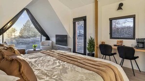 A modern bedroom with a large bed, a desk, two chairs, a small table with a plant, and a TV. - Evergreen Cabin Ocean Front Acadia A Frame Studio (Tremont)