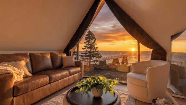 A modern living room with a leather sofa, a white armchair, and a coffee table with a plant. A large window offers a view of the sunset over the ocean. - Sunset Oceanfront Cabin with Sauna, Beach and Dock (Tremont)