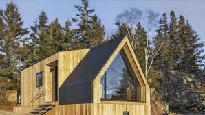 A wooden cabin with a triangular roof, a large window, and a staircase leading to the entrance. - Sunset Oceanfront Cabin with Sauna, Beach and Dock (Tremont)