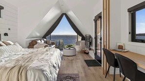 A bedroom with a large bed, a desk, and a view of the sea. - Sunset Oceanfront Cabin with Sauna, Beach and Dock (Tremont)