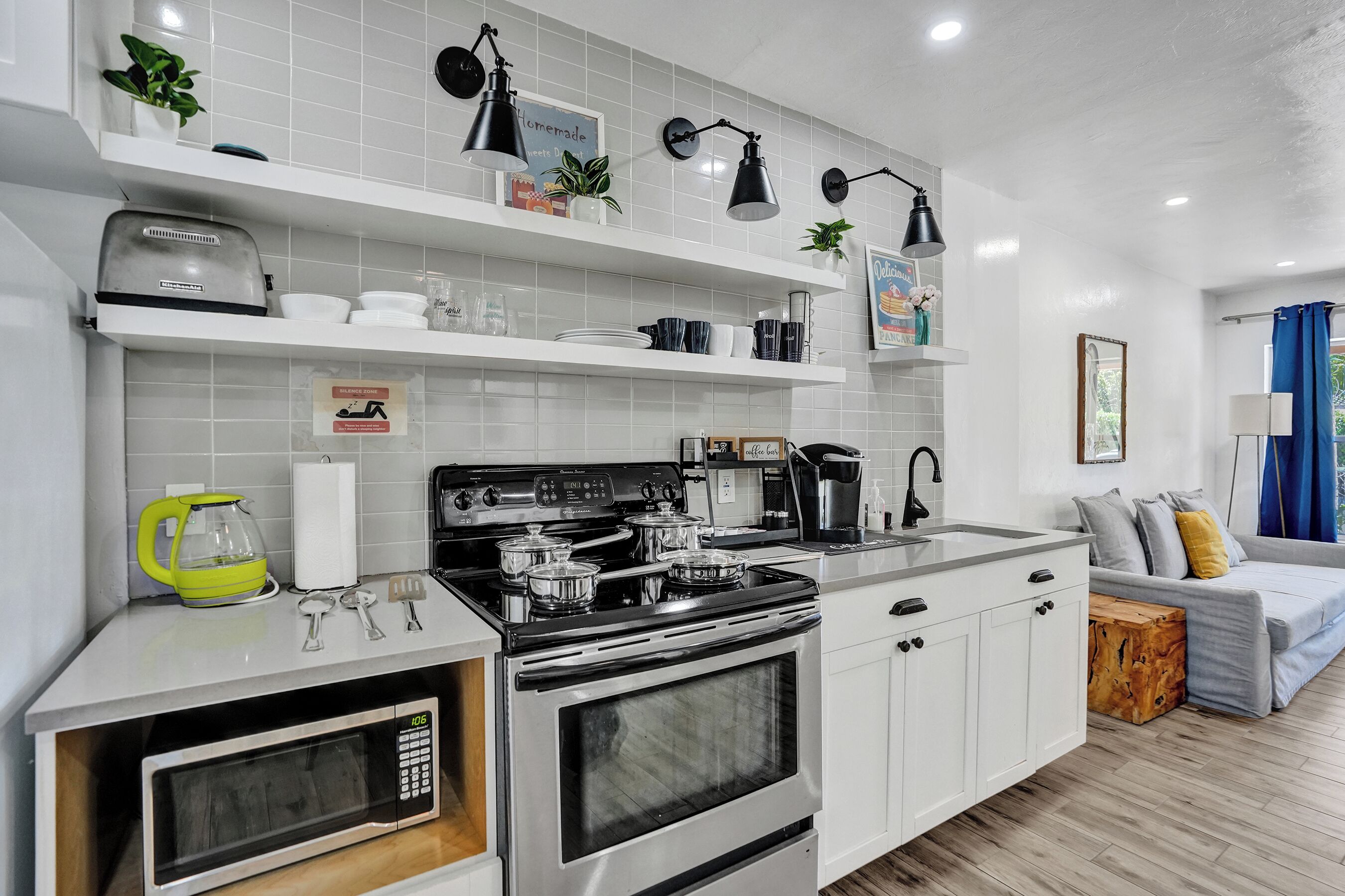 Una cocina moderna con armarios blancos, estufa integrada y microondas. Hay estanterías con plantas y utensilios de cocina, además de una cafetera sobre la encimera.