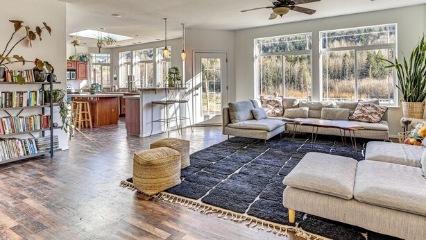 A spacious living area with a large sectional sofa, a coffee table, and a bookshelf filled with books. There is a kitchen area with a bar and a dining table. The room has large windows offering a view of the outdoors. - Nature Art Retreat on Breckenridge Creek w/ Pond! (Sumas)