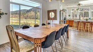 A spacious dining area with a large wooden table, modern chairs, and a view of a mountainous landscape through large windows. - Nature Art Retreat on Breckenridge Creek w/ Pond! (Sumas)