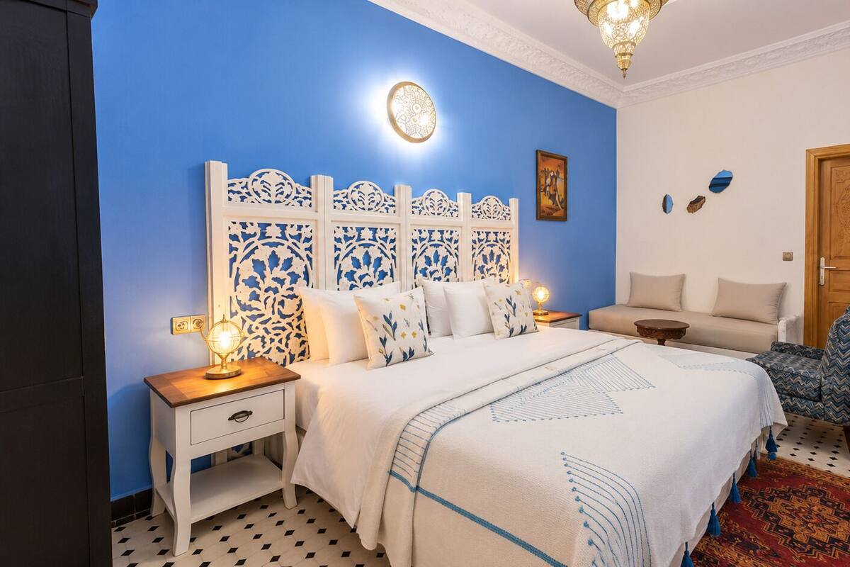 A bedroom with a bed, nightstands, a chair, a sofa, a patterned rug, and a decorative wall panel.