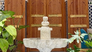 Uma porta de madeira com esculturas detalhadas, uma fonte de mármore e plantas em vasos.