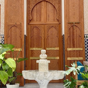 Una puerta de madera con intrincados grabados, una fuente de mármol y plantas en macetas.