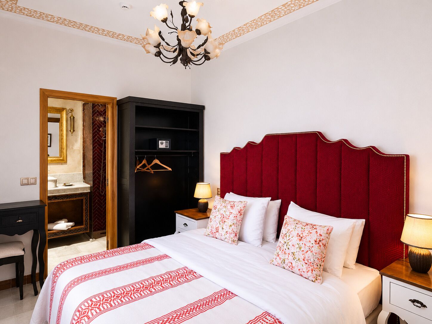 A hotel room with a bed, bedside table, nightstand, wardrobe, desk, and a chandelier.