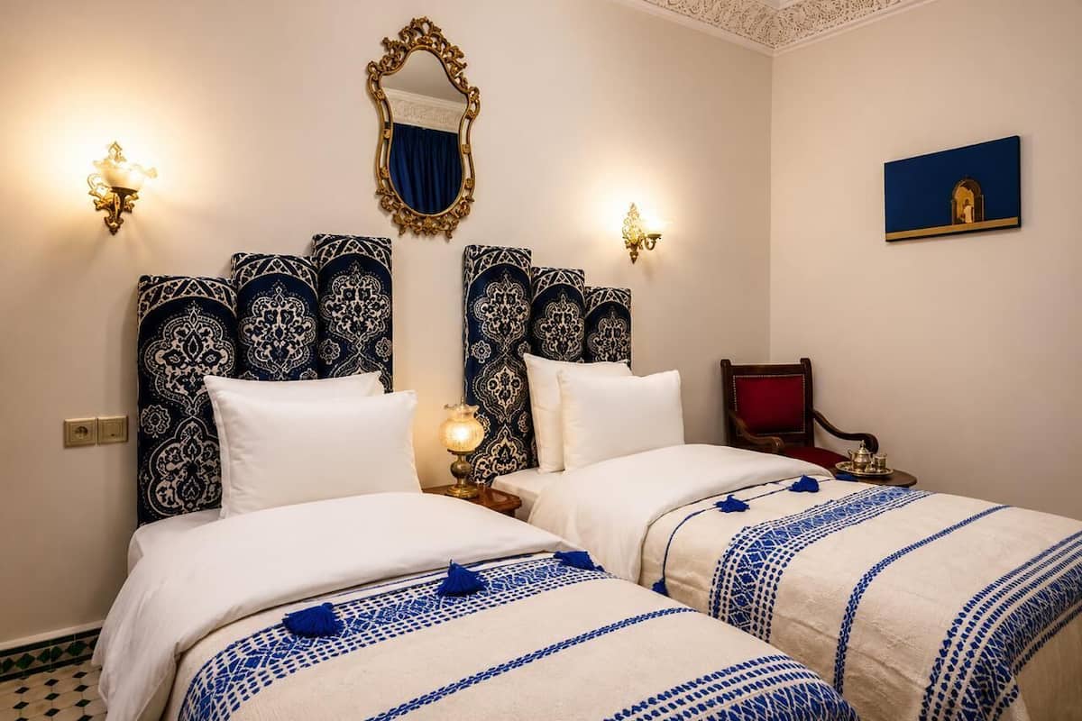 A hotel room with two beds, ornate headboards, wall sconces, a red chair, and a small table with a lamp.
