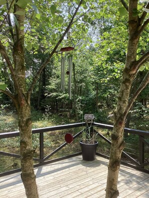 A wooden deck with a railing, surrounded by trees and a hanging wind chime. - Holiday home in the forest close to water (Hadsund)