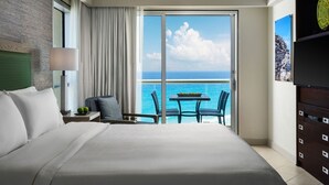 Ein Hotelzimmer mit einem großen Bett, einem Schreibtisch mit Lampe, einem Sessel, einem Tisch mit Obst und einem Balkon mit Meerblick.