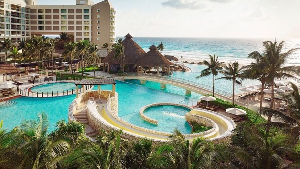 Ein Resort mit einem großen zentralen Pool, einem kleineren Pool und einem Strandbereich mit Sonnenschirmen.