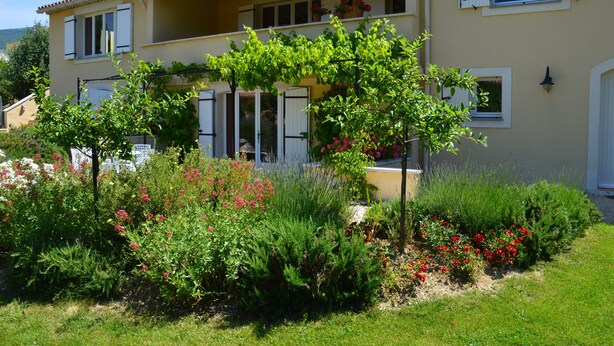 Un edificio de dos pisos con balcón, rodeado por un jardín con diversas plantas y flores.