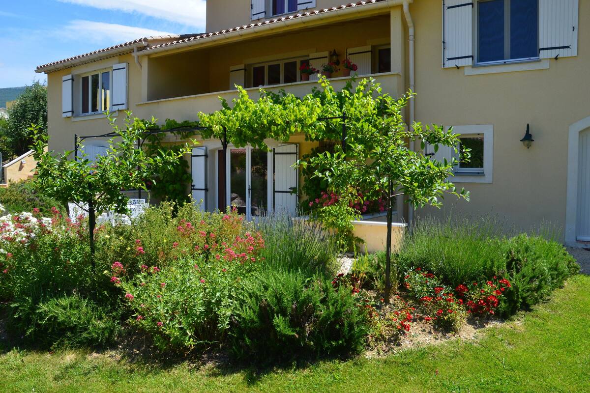 Un edificio de dos pisos con balcón, rodeado de un jardín con diversas plantas y flores.