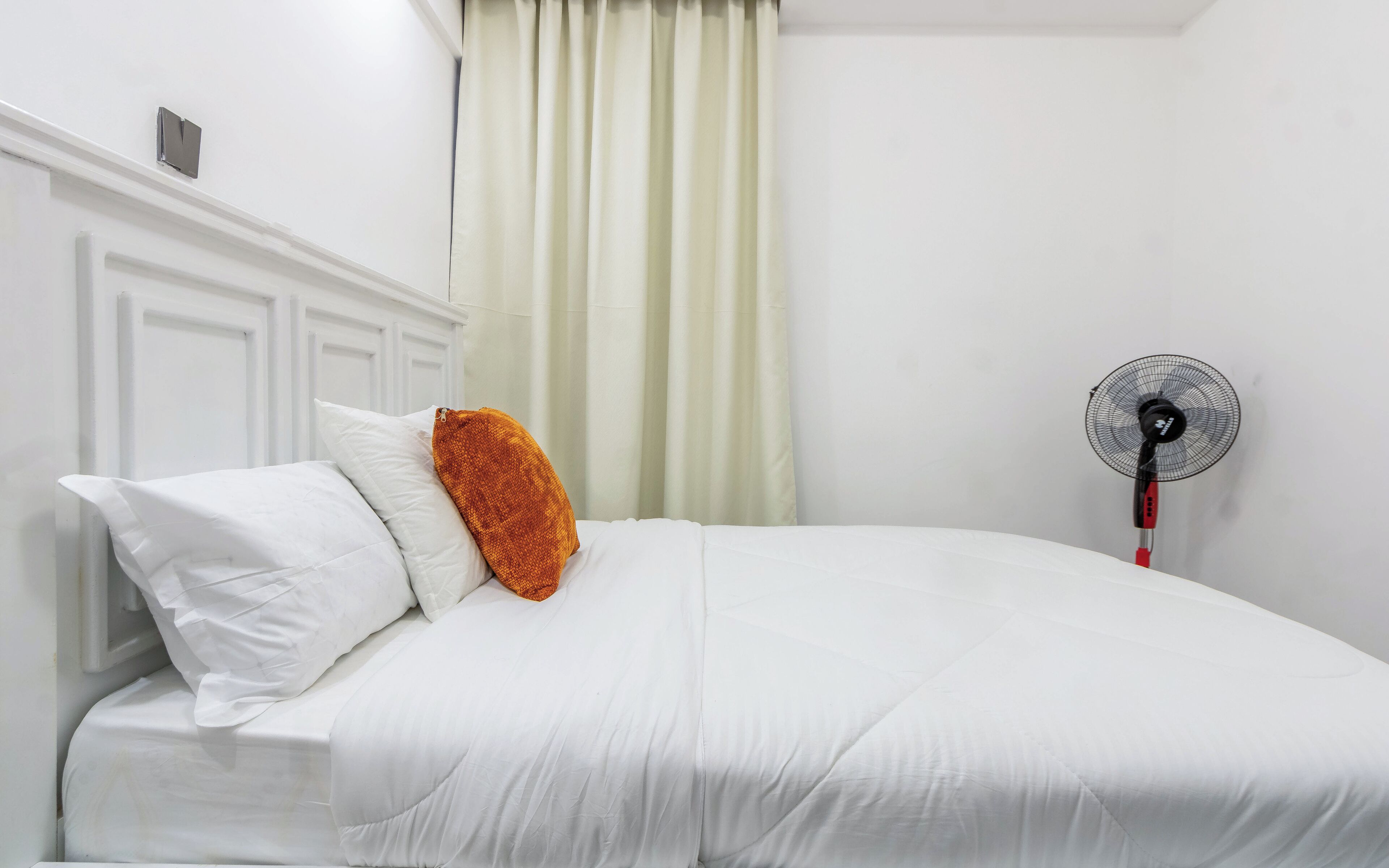 Un lit bien fait, avec une literie blanche et un seul oreiller orange, un mur blanc, une porte blanche et un ventilateur sur pied.