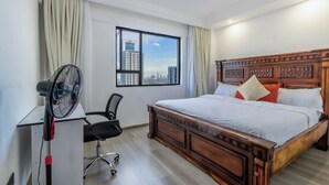 A hotel room with a large bed, a desk with a chair, a standing fan, and a window with a city view. - Shangrila Residency  (Nairobi)