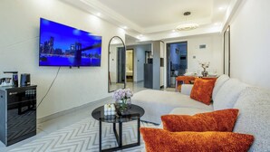 A modern living room with a large flat-screen TV mounted on the wall, a sofa with orange cushions, a coffee table, and a kitchen area visible through an open doorway. - Shangrila Residency  (Nairobi)