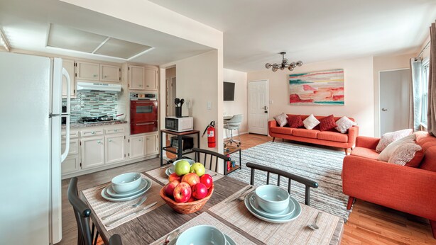 Una cocina moderna con armarios blancos, una mesa de comedor con individuales y un frutero. Hay un sofá rojo en la sala de estar con un cuadro abstracto y colorido en la pared.