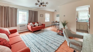 Una sala de estar con un sofá rojo, un sillón gris, una mesa de comedor, y una cocina visible a través de una puerta abierta.
