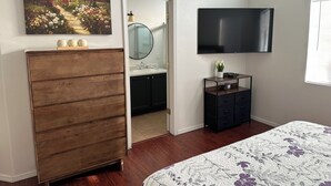 A bedroom with a wooden dresser, a vanity with a mirror, a television mounted on the wall, and a floral bedspread.